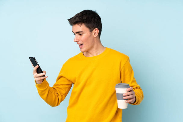Teenager caucasian handsome man isolated on purple background holding coffee to take away and a mobile - Photo, Image