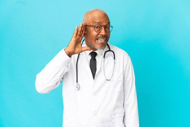 Senior arts man geïsoleerd op blauwe achtergrond luisteren naar iets door het leggen van de hand op het oor - Foto, afbeelding