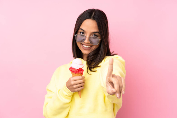 Giovane ragazza bruna che tiene un gelato alla cornetta su uno sfondo rosa isolato che mostra e solleva un dito - Foto, immagini