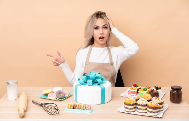 Teenager pastry chef with a big cake in a table surprised and pointing finger to the side - Photo, image