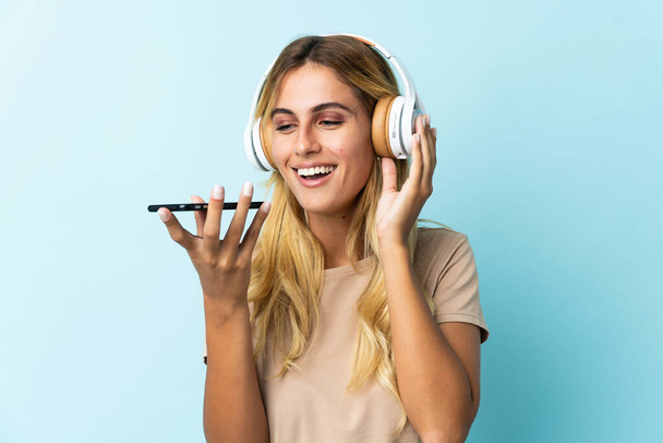 Молода блондинка Уругвай ізольована на синьому фоні, слухаючи музику мобільним телефоном і співаючи - Фото, зображення