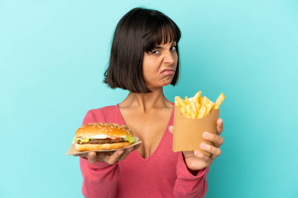 Молодая брюнетка с гамбургером и жареными чипсами на изолированном синем фоне - Фото, изображение