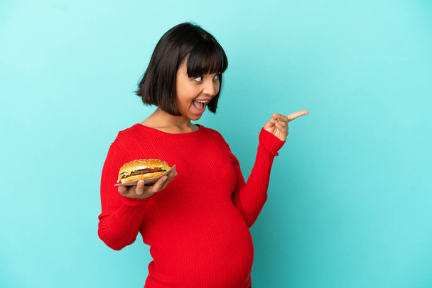 Mujer embarazada joven sosteniendo una hamburguesa sobre un fondo aislado con la intención de darse cuenta de la solución mientras levanta un dedo - Foto, imagen