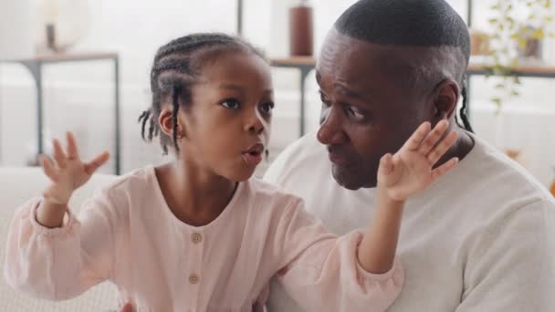 Afro amerykański Dojrzały mężczyzna Afryki ojciec czarny dziadek siedzi na kanapie w domu z małą dziewczynką cute córka dziecko rozmowy rozmowy, dziecko wyjaśniające wiadomości akcji, komunikacji rodzinnej - Materiał filmowy, wideo