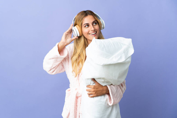 Young blonde Uruguayan woman isolated on blue background in pajamas and holding a pillow and listening music - Fotoğraf, Görsel
