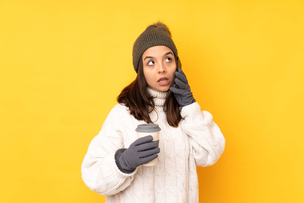 Jeune femme avec chapeau d'hiver sur fond jaune isolé tenant café à emporter et un mobile - Photo, image