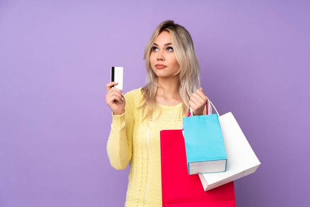 Teenager blonde girl over isolated purple background holding shopping bags and a credit card and thinking - Foto, imagen