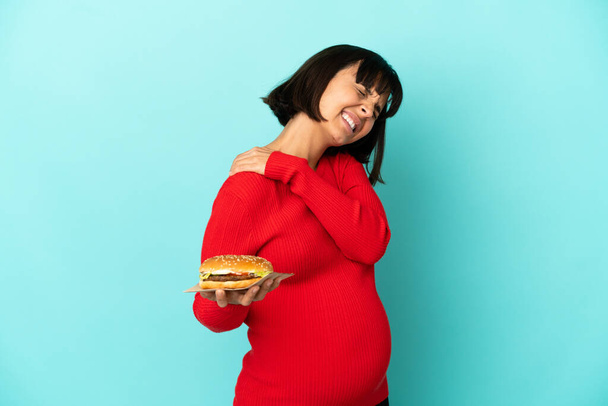 Молода вагітна жінка тримає бургер над ізольованим тлом, що страждає від болю в плечі за зусилля
 - Фото, зображення