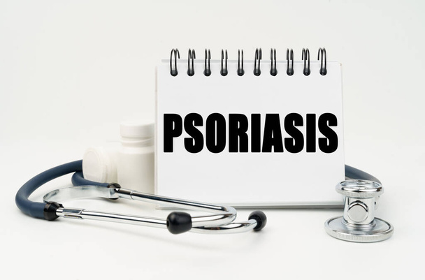 Medicine and health concept. On a white table lies a stethoscope, pills and a notebook with the inscription - PSORIASIS - Фото, изображение
