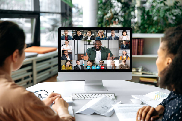 Virtual communication, video call. View over the shoulders of two women to a computer screen with successful multiracial business team gathered in a video conference to discuss working issues,strategy - Photo, Image