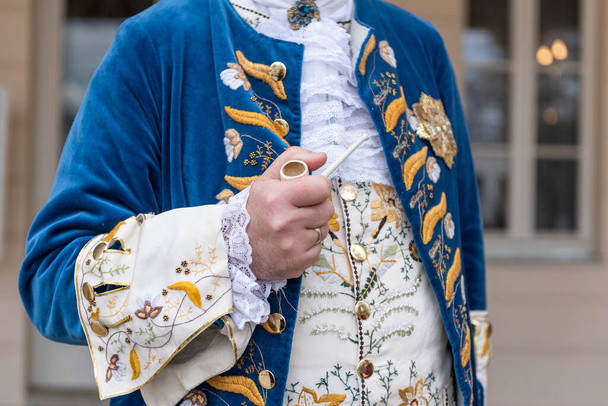 Die Details eines Mannes in einem barocken Kostüm. Eine Hand hält eine Pfeife, goldene Knöpfe, Weste und dekorative Säume. - Foto, Bild