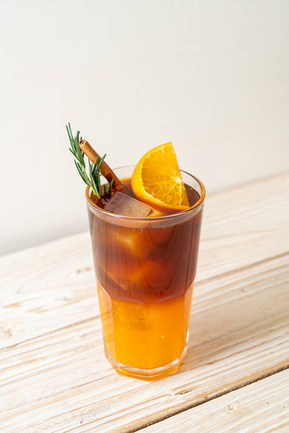 A glass of iced americano black coffee and layer of orange and lemon juice decorated with rosemary and cinnamon on wood background - Foto, imagen