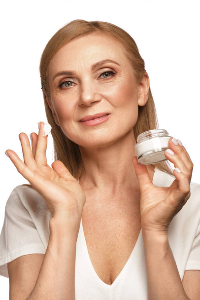 Portrait of a beautiful elderly woman in a white shirt with a moisturizing face cream in her hands. - Zdjęcie, obraz