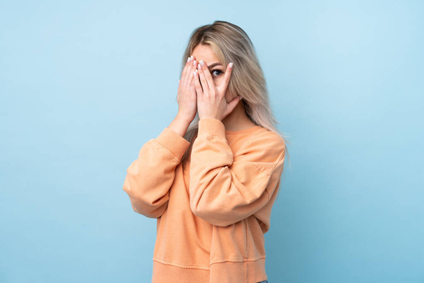 tiener meisje over geïsoleerde blauwe achtergrond bedekken ogen en kijken door vingers - Foto, afbeelding