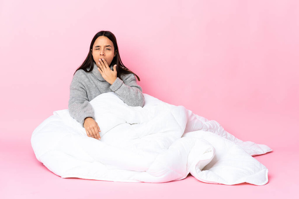 Молодая женщина смешанной расы в пижаме сидит на полу зевая и прикрывая широко открытый рот рукой - Фото, изображение