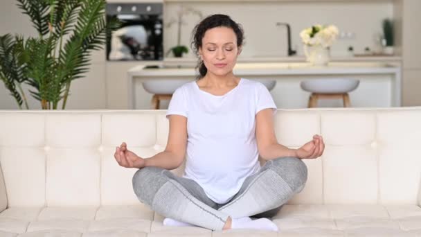Calm beautiful happy pregnant mixed race woman, resting while sitting at home in living room on sofa in lotus position, eyes closed, smiling. The expectant mother is chilling, relieves stress - Footage, Video