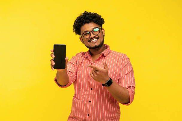 Smiling indian guy isolated on yellow background holding smartphone with black screen in hand and pointing finger at it, looking at camera, place for text. Studio photo. - Foto, afbeelding