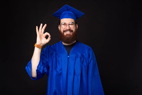 gelukkig jong student in bachelor staan over zwart achtergrond en tonen ok gebaar. - Foto, afbeelding