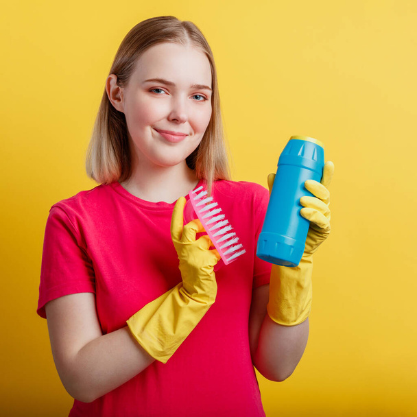 Junge blonde fröhlich lächelnde Frau bereit, Haus mit Hausrat isoliert über gelbem Hintergrund zu putzen. Frauenporträt mit Reinigungsbürste in Gummihandschuhen. Nahaufnahme Platz - Foto, Bild