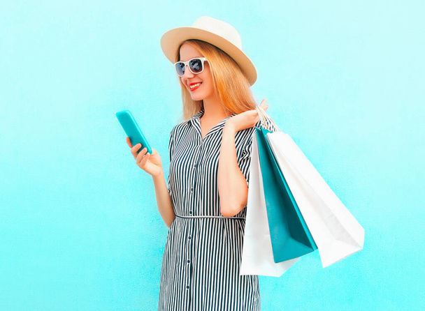 Portrait of happy smiling young woman with smartphone and shopping bags wearing a summer straw hat, striped dress on a blue background - Zdjęcie, obraz