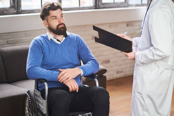 Handsome bearded man sitting in a wheelchair and listening to a doctor - Fotoğraf, Görsel