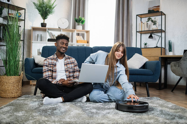 Couple using laptop while robot vacuum cleaning carpet - 写真・画像