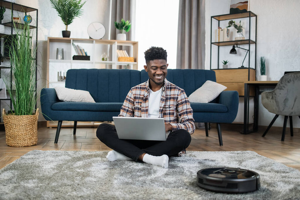 Afro człowiek za pomocą laptopa podczas odkurzacza robi prace domowe - Zdjęcie, obraz