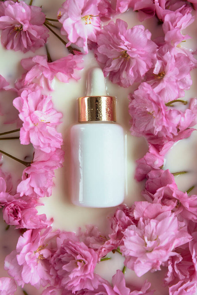 White glass bottle of cosmetic serum on a background of pink spring flowers in water or milk. Natural organic cosmetics concept. Top view, flat lay, mock up - Photo, Image