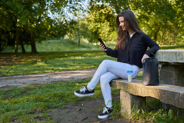 hosszú hajú, fekete inges, fehér nadrágos lány, aki a telefonját nézegeti a természetben, egy kőasztalnál ül egy kávéval és a táskájával. nő beszélget mobil, miközben pihen a természetben. tini a parkban - Fotó, kép
