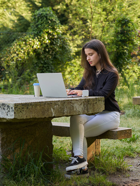 hosszú hajú, csinos üzletasszony laptoppal, fejhallgatóval és kávéval a kőasztalnál. tinédzser dolgozik a laptopján a természetben, fókuszálatlan zöld háttérrel. lány számítógép szabadban. - Fotó, kép