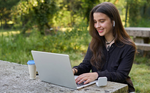 long-haired pretty businesswoman with laptop, headphones and coffee at stone table. teenager working on her laptop in nature with unfocused green background. girl with computer outdoors. - Photo, Image