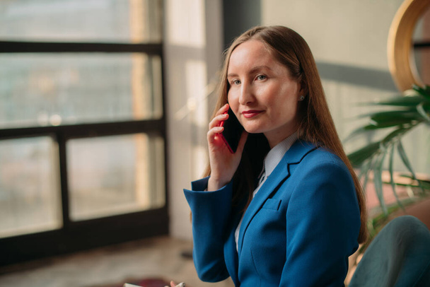 現代オフィスインテリアのビジネス女性の肖像画。携帯電話で話して青いビジネススーツのスタイリッシュな若い女の子。ビジネスにおける女性、ビジネスにおける女性の肖像. - 写真・画像