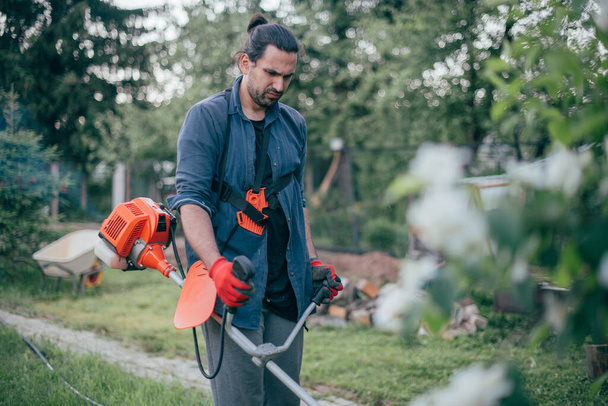 Mies leikkaa nurmikon puutarhassa ruohonleikkurilla. Nuori kaveri huolehtii nurmikon ja työskentelee puutarhassa - Valokuva, kuva