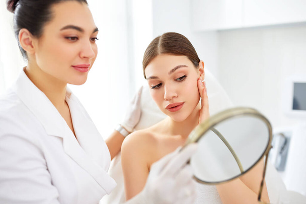 Elégedett fiatal nő néz tükör és megható arc után professzionális kozmetikai kezelés kozmetikus a modern kozmetikai klinika - Fotó, kép