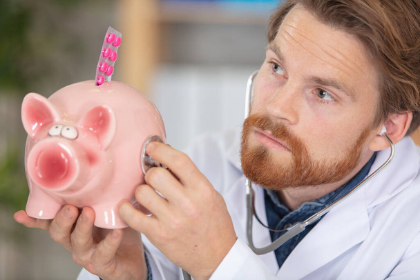 doctor holding stethoscope to piggybank continaing blister of tablets - Foto, Bild