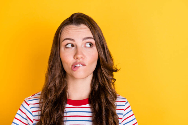 Photo of sweet doubtful young woman wear striped outfit biting lip looking empty space isolated yellow color background - 写真・画像