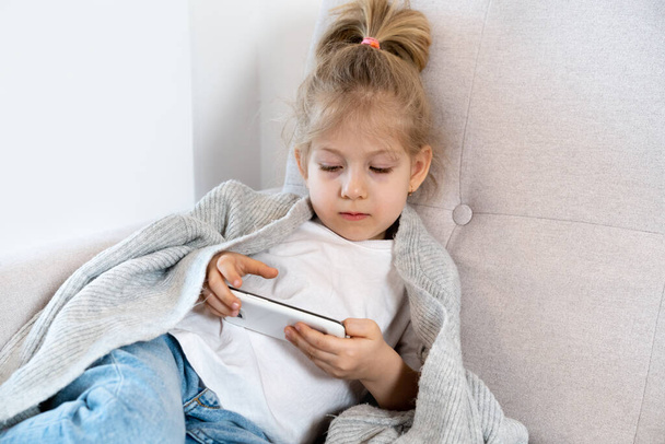 Little beautiful blonde girl on the couch with phone in her hands. the concept of modern childhood. - Fotó, kép