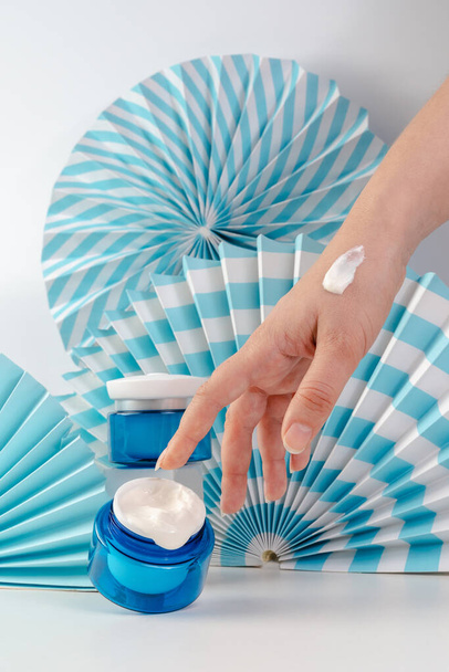 Νεαρή γυναίκα κρατά ένα βάζο με κρέμα προσώπου στα χέρια της. Αντιρυτιδική κρέμα σε μπλε φόντο. Αντιγηραντικά καλλυντικά για άνδρες και γυναίκες. Προϊόντα Unisex. Mockup. Ιάπωνες θαυμαστές. Θεραπείες Spa για το δέρμα  - Φωτογραφία, εικόνα