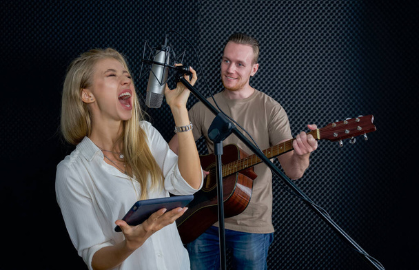 Young female singing with tablet computer in her hand. While the man playing an acoustic guitar in front of black soundproofing walls. Musicians producing music in professional recording studio. - Photo, Image