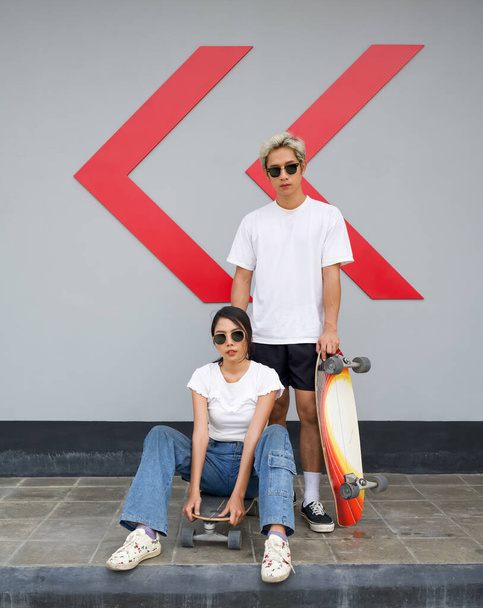 Asian couple resting after skateboarding. A young man in a white T-shirt held his hand on the surfskates board while his girlfriend with sunglasses sit in front. - Foto, Imagem