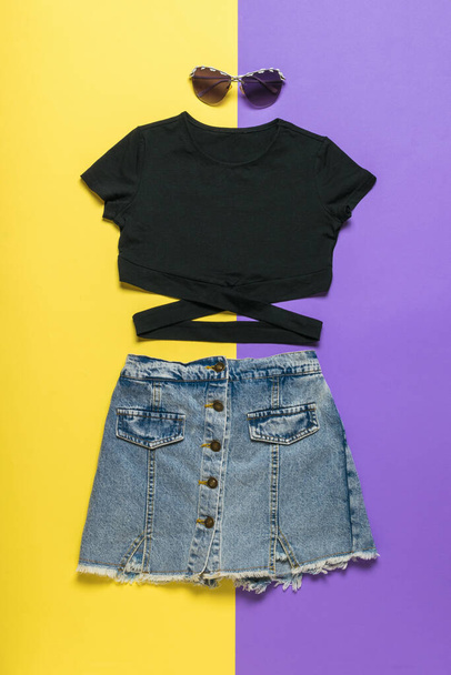 イエローとパープルを基調としたブラックのTシャツ、ブラックのメガネ、デニムのスカート。夏服のセット. - 写真・画像