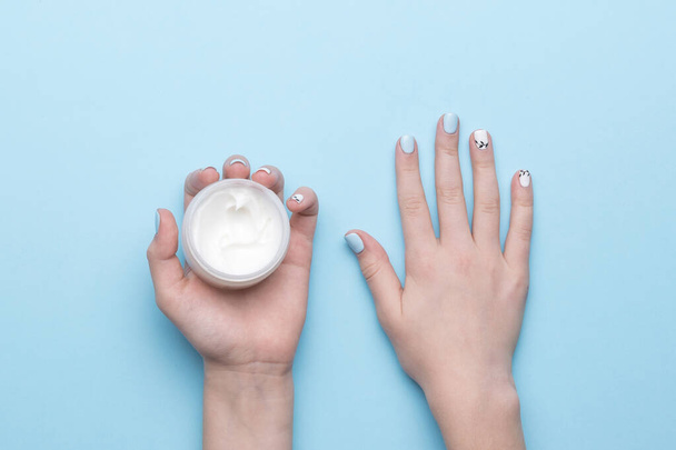 Un frasco de crema en las manos de las mujeres con una manicura elegante sobre un fondo azul. Cuidado de la piel de las manos. Puesta plana. - Foto, Imagen