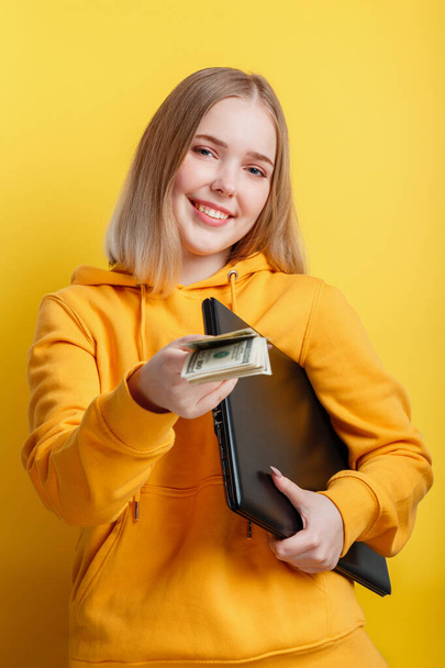 Beautiful smiling young woman freelancer with laptop take dollars money banknotes. IT remote freelancer remote worker via laptop isolated over yellow color background. - Foto, Imagen