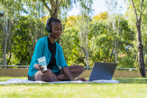 fekete afrikai nő ül egy takarón a parkban használja a számítógépet és a vezeték nélküli headset, miközben kezében egy forró ital - Fotó, kép