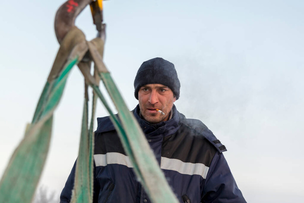 Portrait of a worker in a blue jacket and knitted hat at the hook of a truck crane with a ciga-rette in his mouth - Foto, immagini