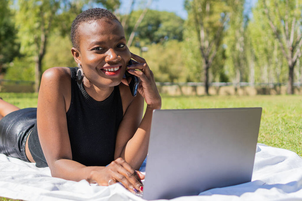 donna africana nera sdraiata su una coperta nel parco a parlare al telefono cellulare, con il computer, guardando dritto nella fotocamera - Foto, immagini
