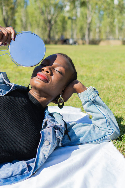black afro-american woman in casual clothes holding a mirror lying on a white blanket, eyes closed, relaxing expression, vertical photo - Photo, Image