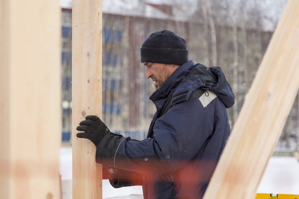 An installer in a hat with earflaps mounts the frame of a slide made of wooden beams - Photo, Image