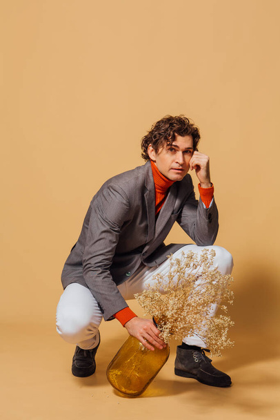 Fashion portrait of a tall handsome man dressed in orange turtleneck, white jeans and grey jacket sitting and holding vase with dry flowers on the beige background - Foto, imagen