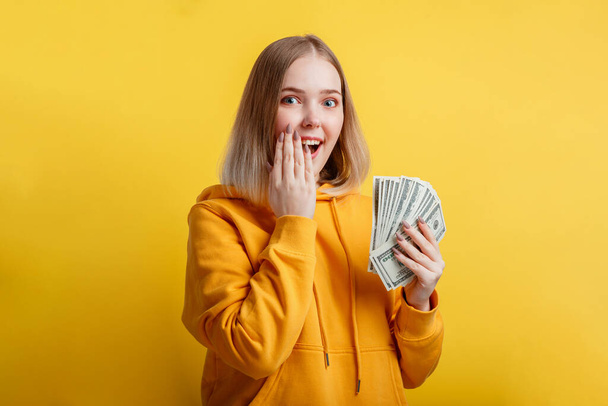 Happy excited rich young woman win money cash. Lady hold Dollar stack surprised rejoices at lottery victory. Teen girl covered her open mouth with hand surprise isolated over yellow color background - Foto, Imagen